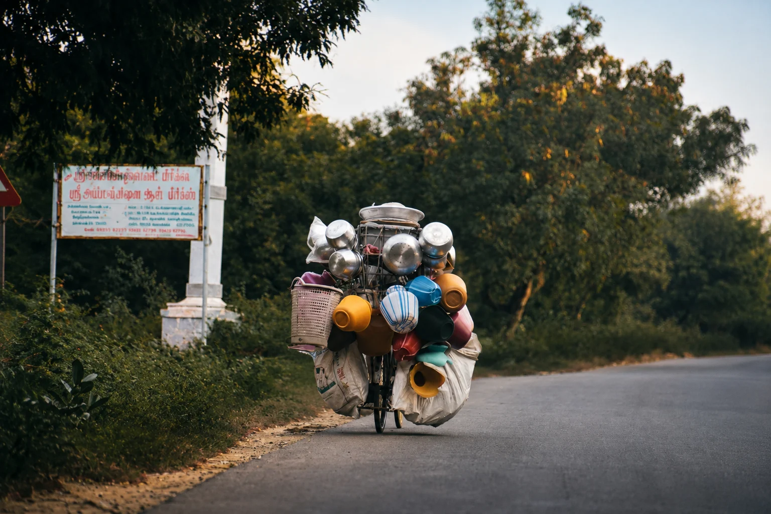 A person travelling along a quiet road on a heavily loaded bike carrying pots, containers and bags.