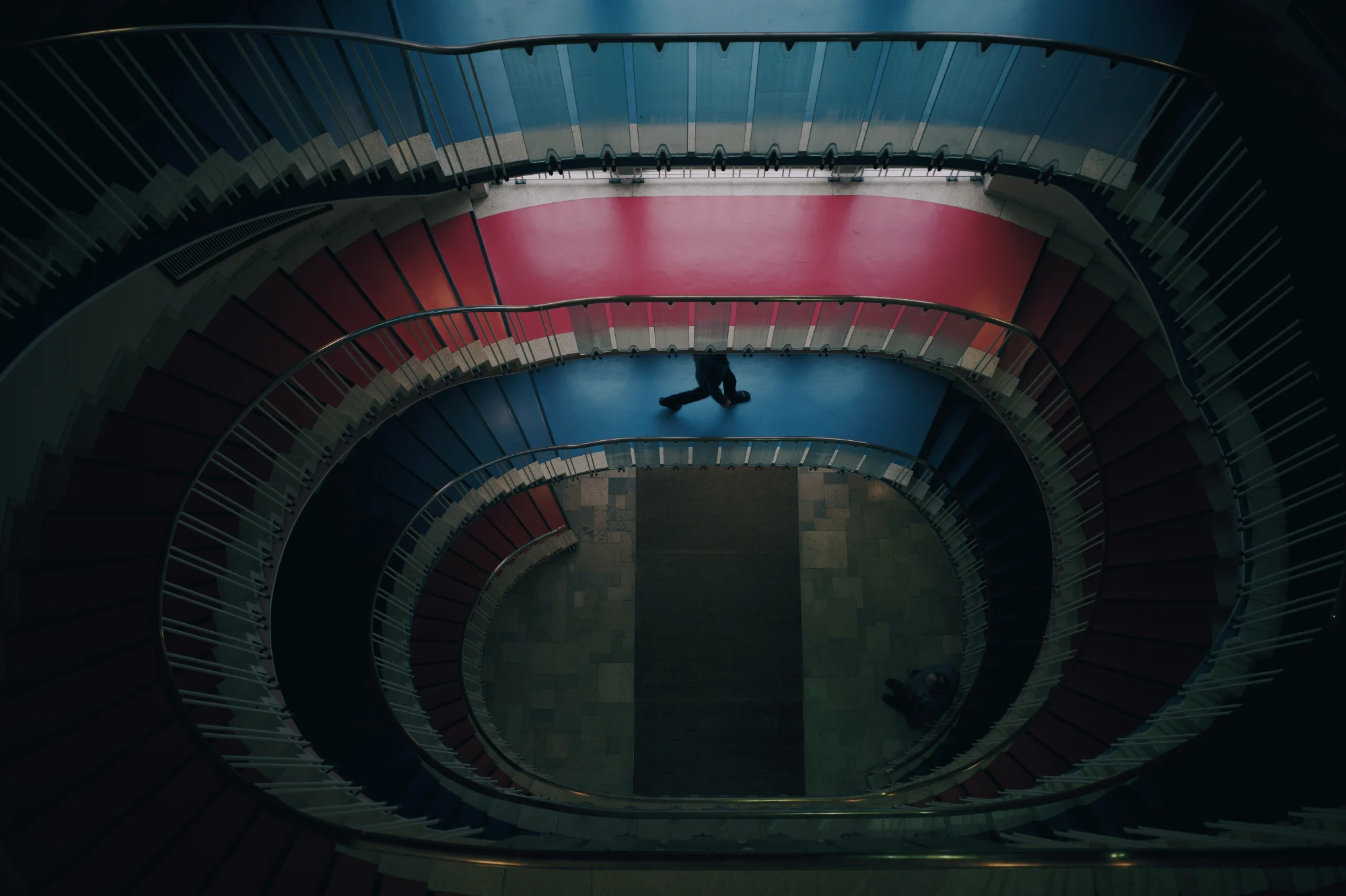 An overhead view of a spiral staircase with red and blue levels, with two people visible on different parts of the staircase.
